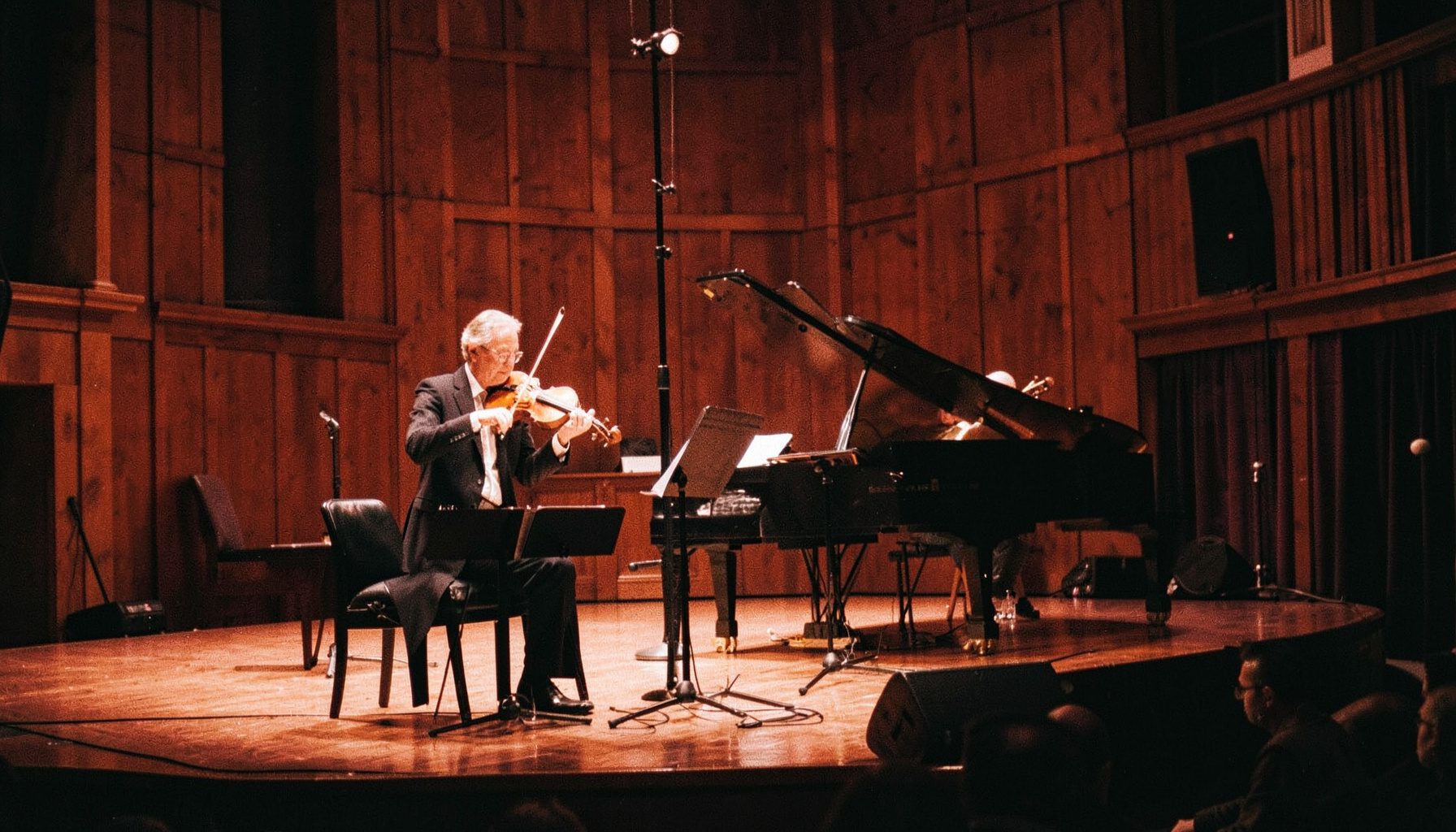 Warmly lit concert stage with violin and piano in a wood-paneled hall, rich amber and deep brown tones